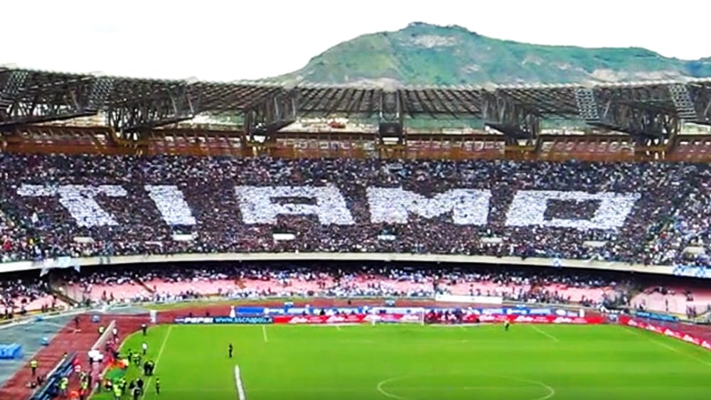Coreografia dei tifosi del Napoli allo stadio: le tribune formano la scritta gigante "TI AMO" in bianco su sfondo azzurro, con il Vesuvio sullo sfondo.